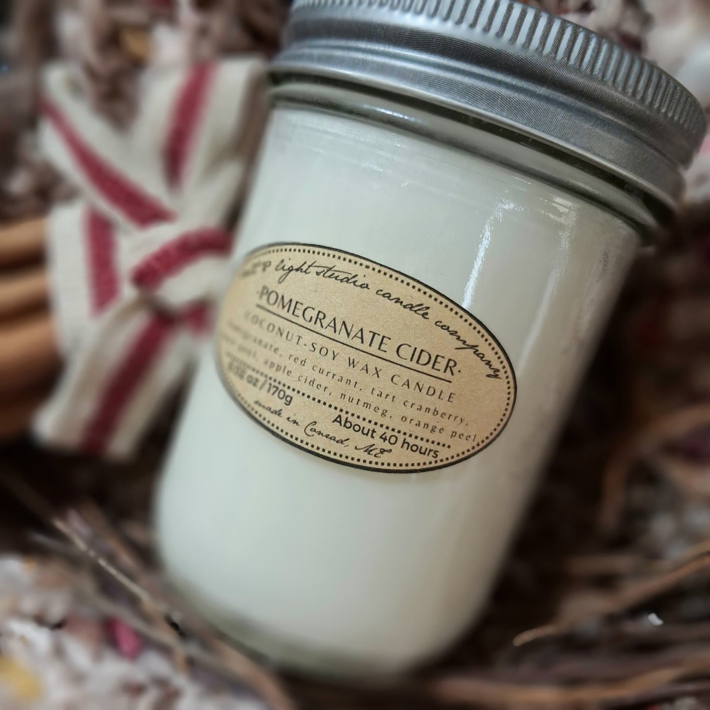 Hand poured candle jar labeled 'Pomegranate Cider' with a bundle of cinnamon sticks wrapped with decorative ribbon in the background