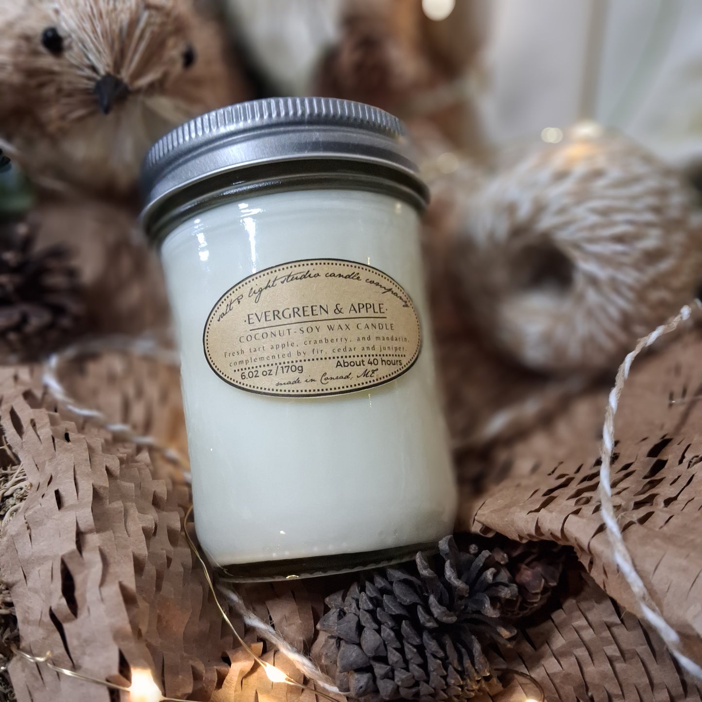 Coconut soy candle evergreen and apple scented with a brown paper label on decorative items including pinecones, cotton twine and string lights