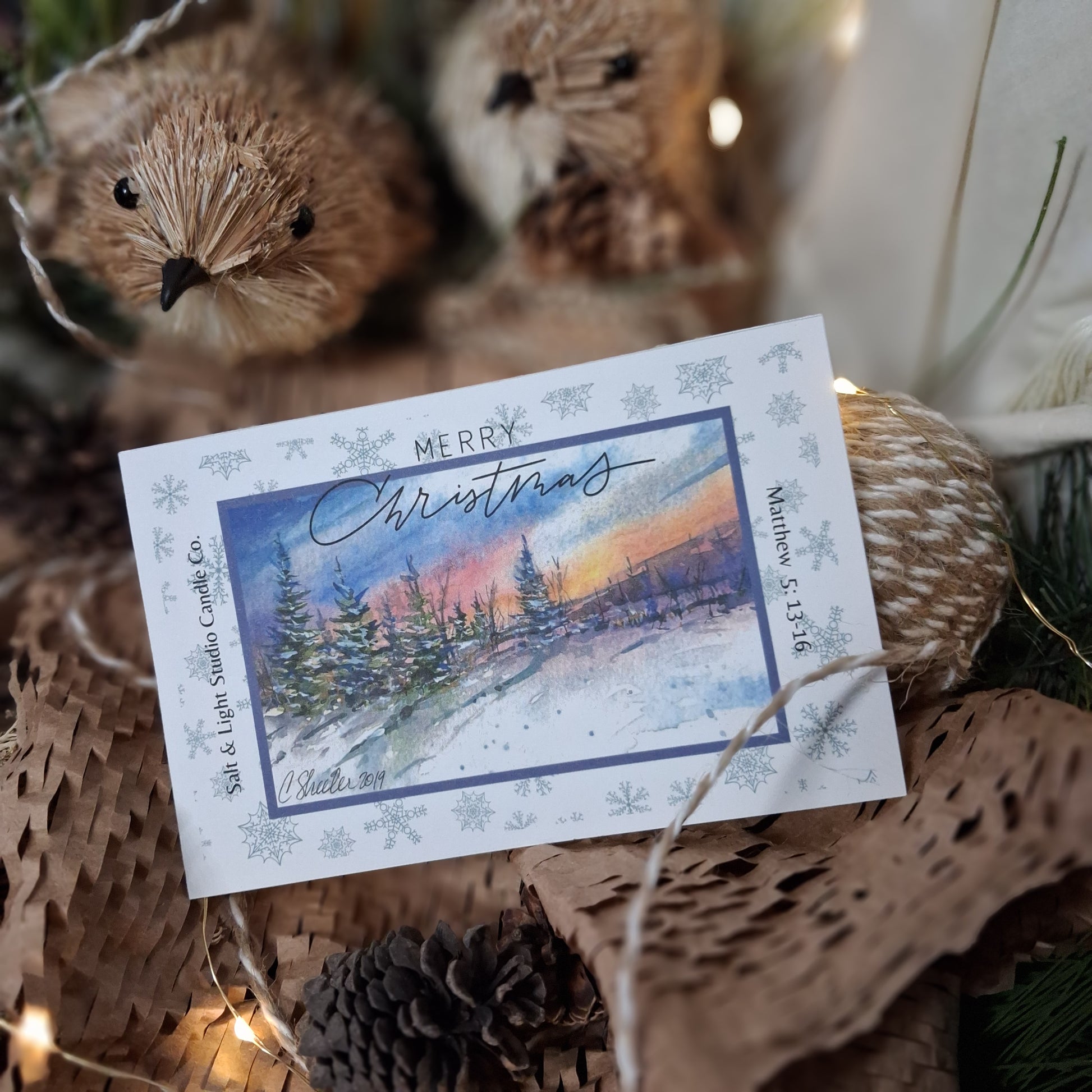 Christmas card with snowy landscape and 'Merry Christmas' text, surrounded by pinecones and birds made of wood.
